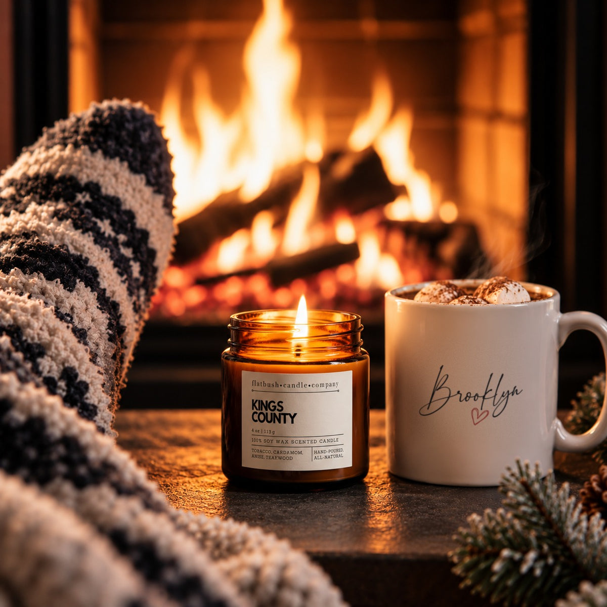 The Night Light gift set: Brooklyn Heart mug of hot cocoa beside a lit Kings County 4 oz soy candle by a fireplace